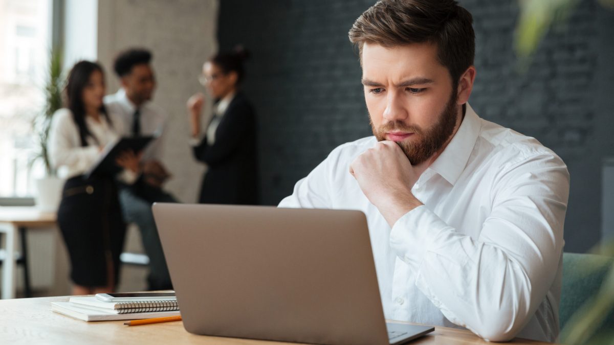 Image of concentrated young caucasian businessman sitting indoors using laptop computer. Looking aside.