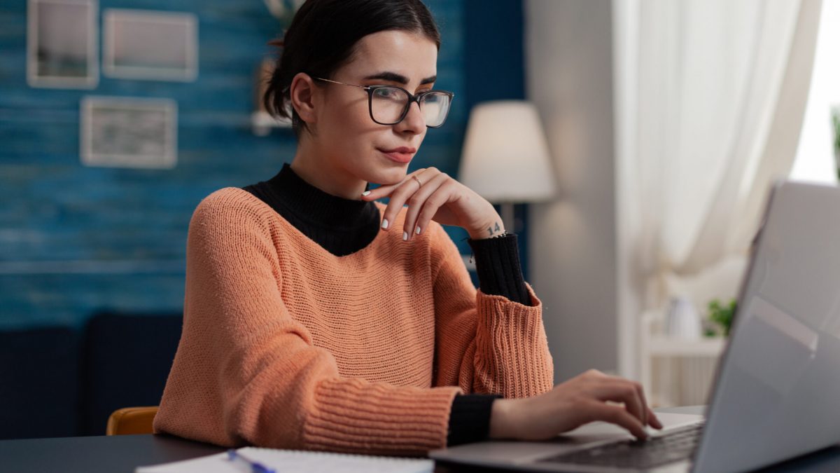 Confident student studying remote looking at laptop screen. Content creator with glasses casually typing sitting at home desk. College girl browsing the internet. Freelancer working from home sending email.